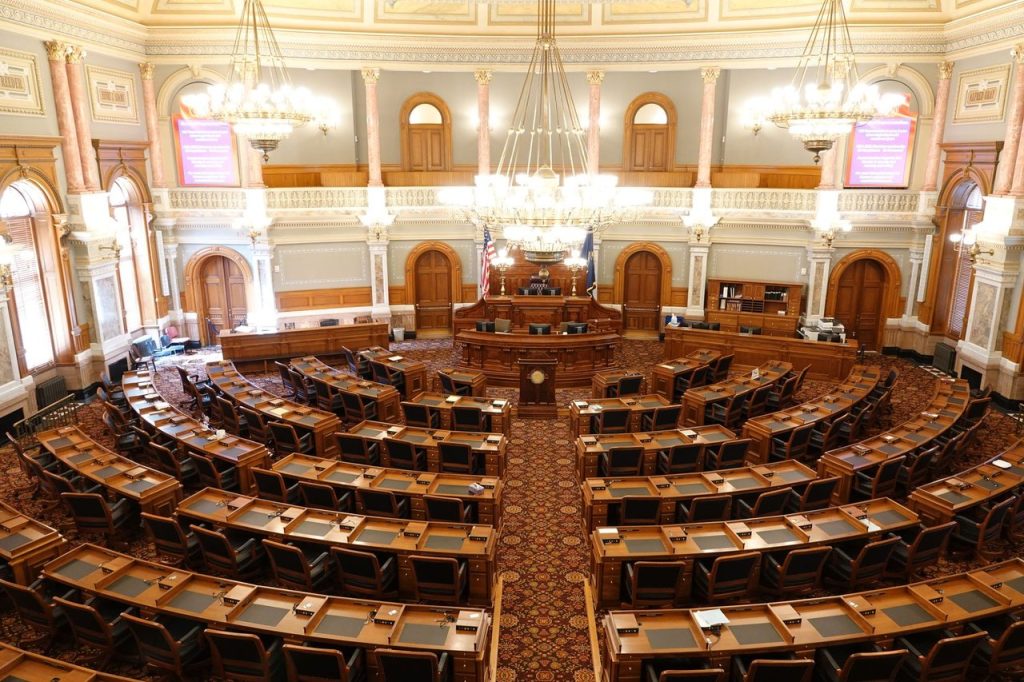 Vista ampla de um plenário legislativo ornamentado, com cadeiras vazias e lustres.