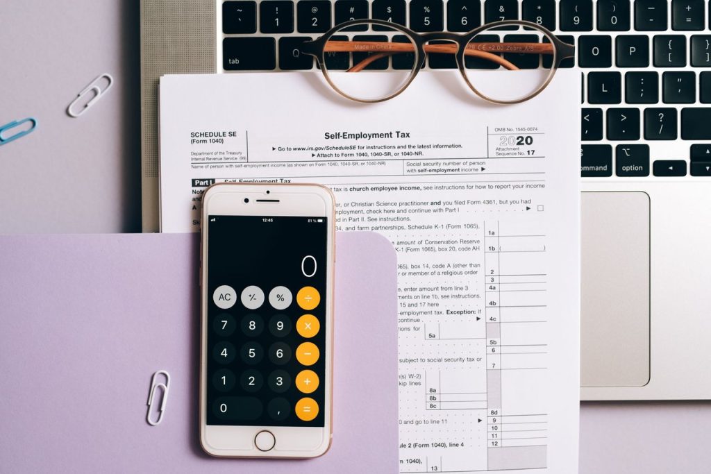 Documentos fiscais, calculadora no celular e laptop organizados sobre uma mesa.