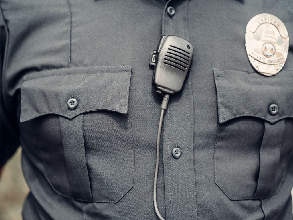Detalhe do uniforme de um policial, com câmera corporal e distintivo visíveis.