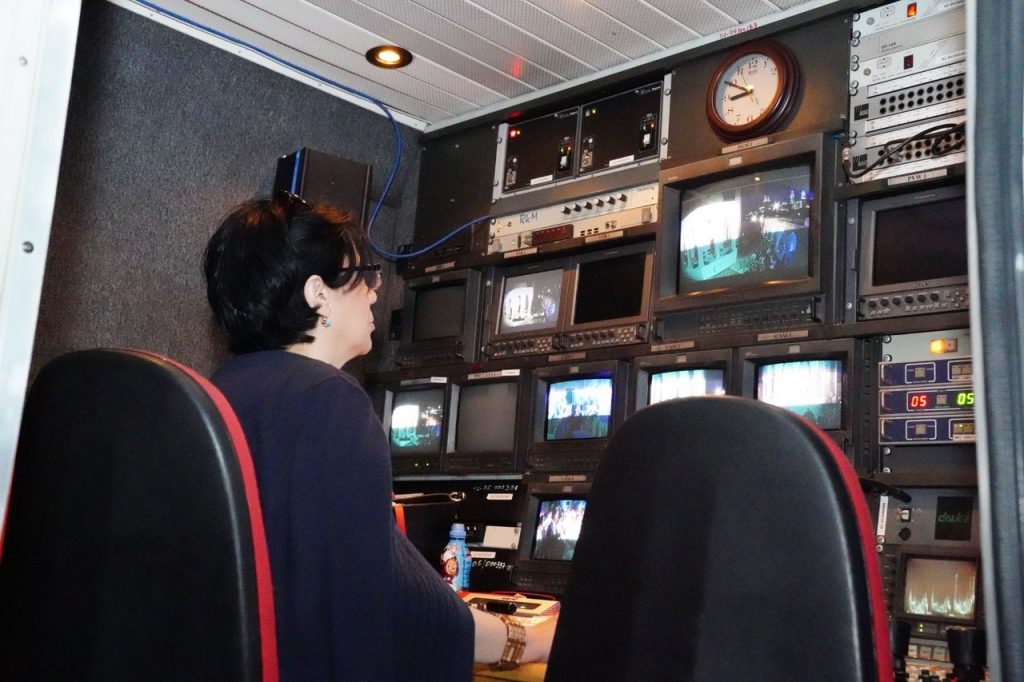 Profissional opera equipamentos em uma sala de controle de televisão com várias telas e dispositivos de transmissão.