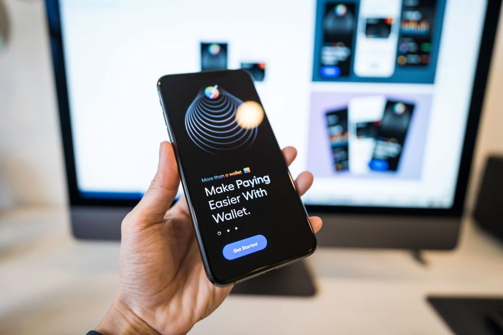Mão segura um smartphone com a tela de uma carteira digital aberta, diante de um monitor desfocado.